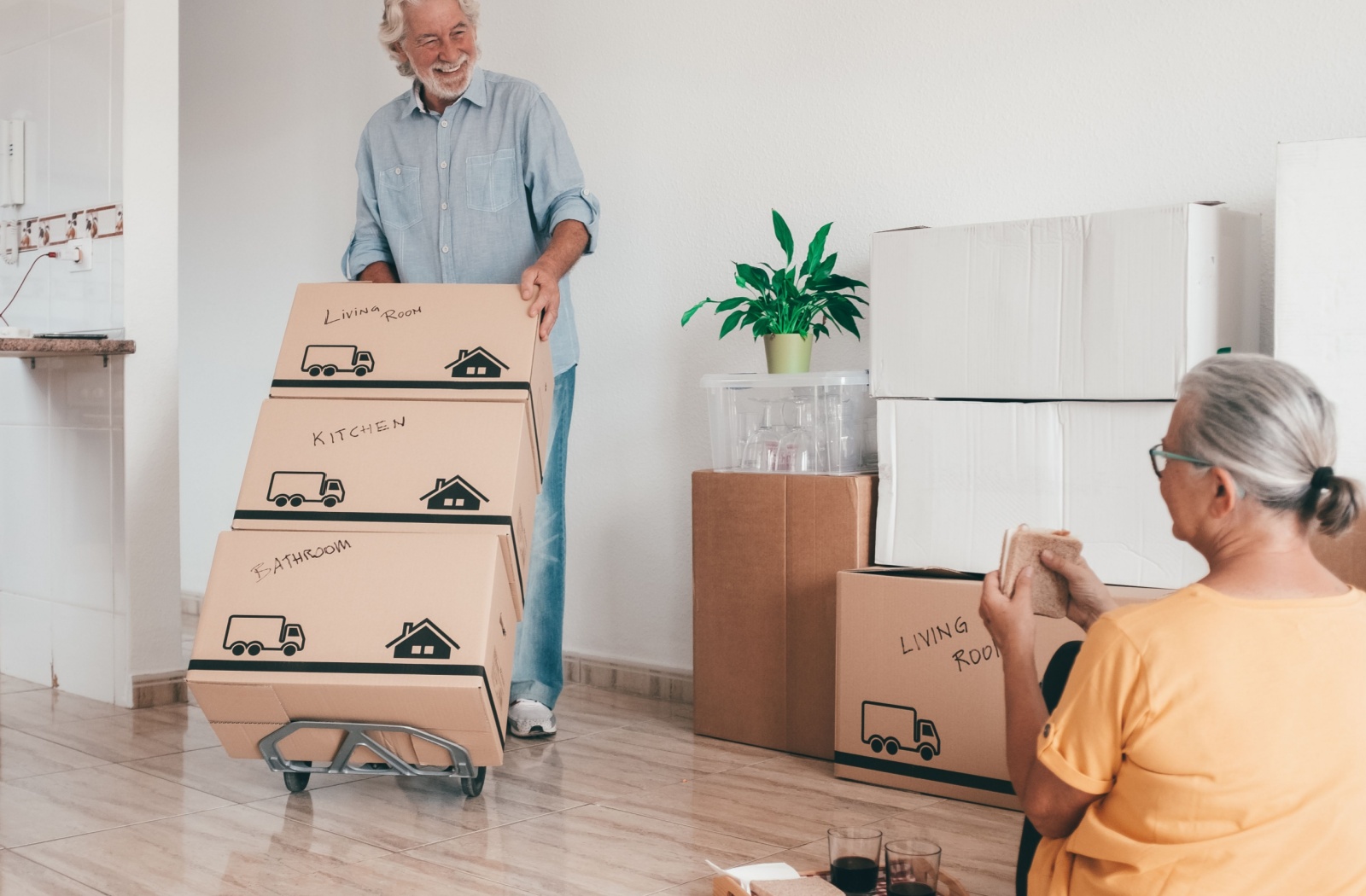 A man happily moves labeled boxes while a woman sits nearby, enjoying a snack.