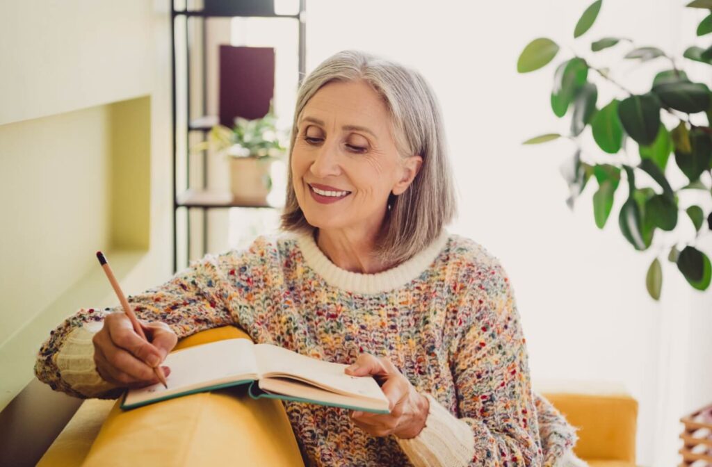 A smiling older adult writing in a notebook with a pencil to plan out their morning.