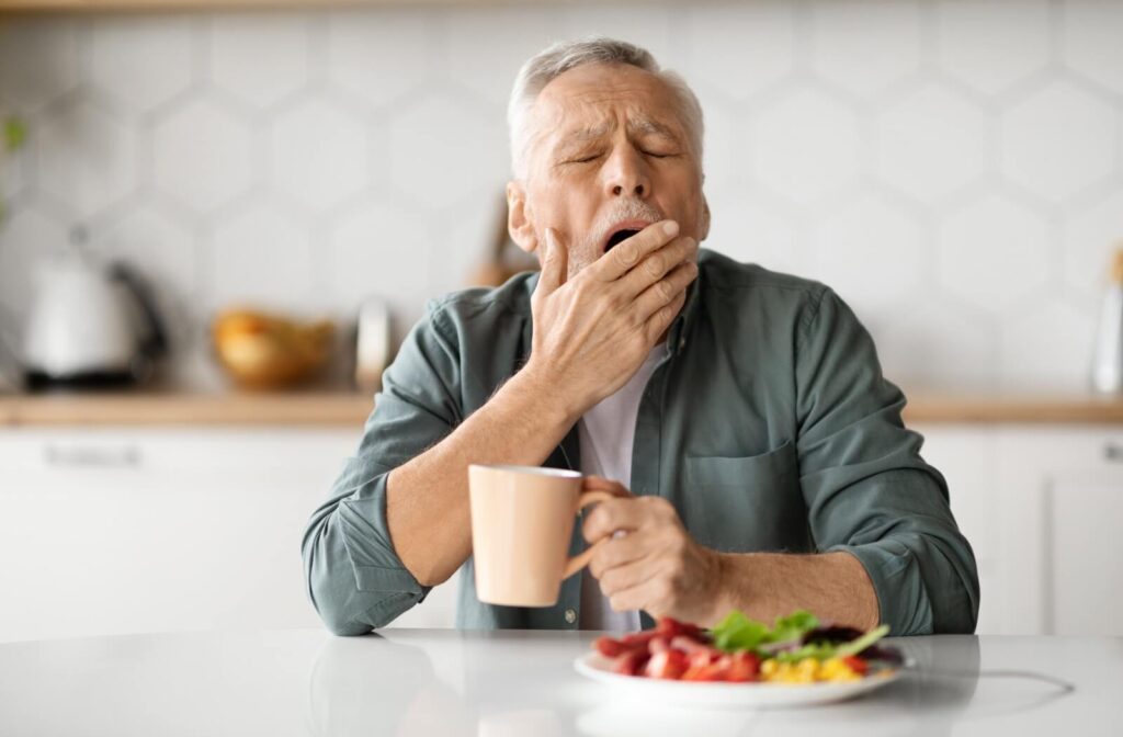 An older adult yawns over a colorful breakfast plate while drinking coffee in their kitchen to combat the symptoms of their chronic fatigue